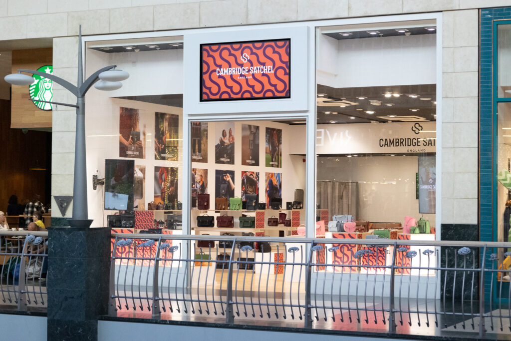 Exterior of a Cambridge Satchel store in a mall with displayed bags and a Starbucks sign nearby.