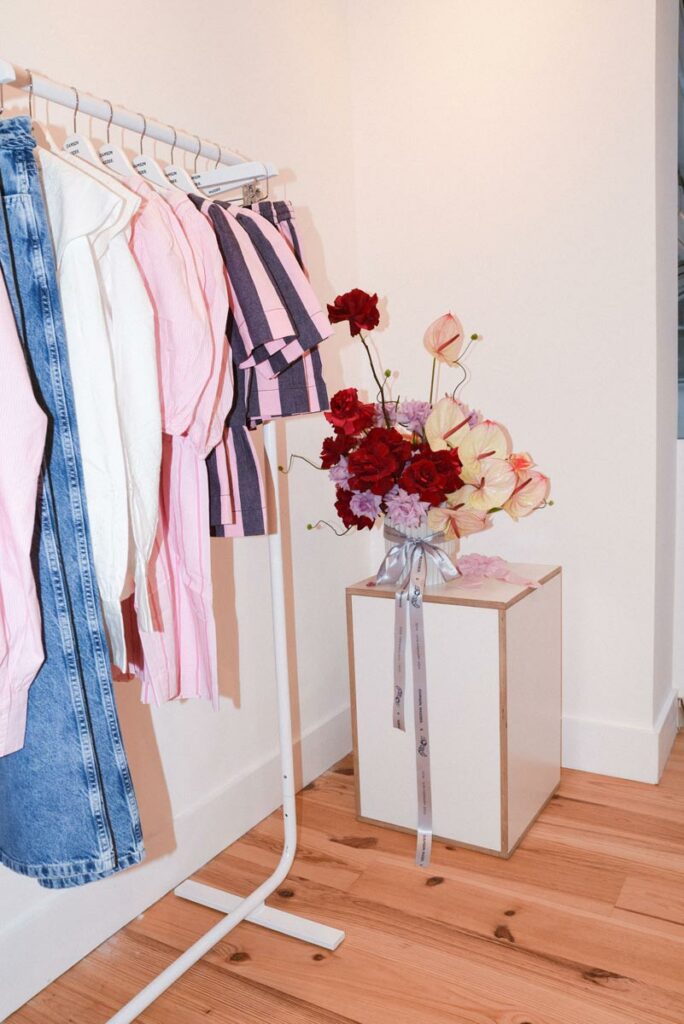 Fashion clothing on a rack and a bouquet of red flowers on a pedestal in a well-lit room.