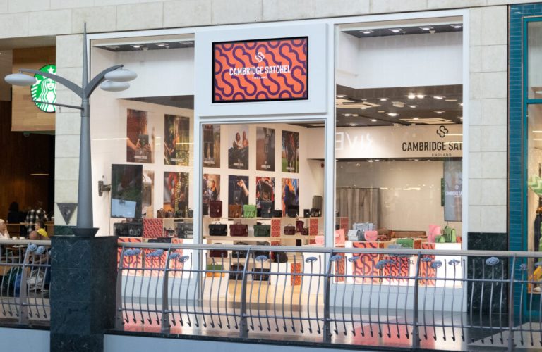 Exterior of a Cambridge Satchel store in a mall with displayed bags and a Starbucks sign nearby.